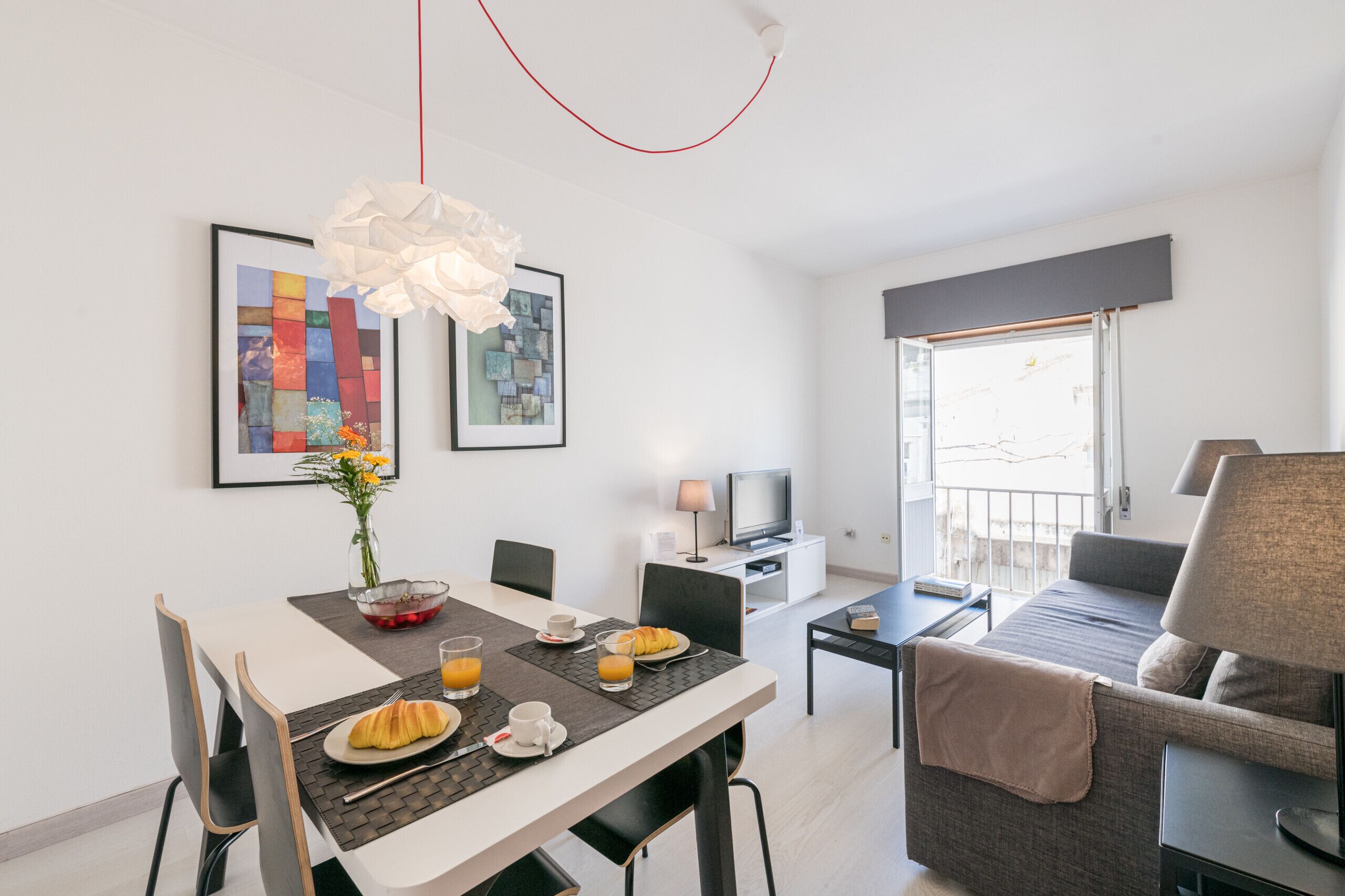 A modern hotel room with a dining area, a sofa, a TV, and artwork on the walls.
