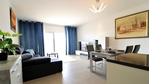 Een moderne woonkamer met een bank, een eettafel gedekt voor twee personen, een televisie en een groot schilderij aan de muur.