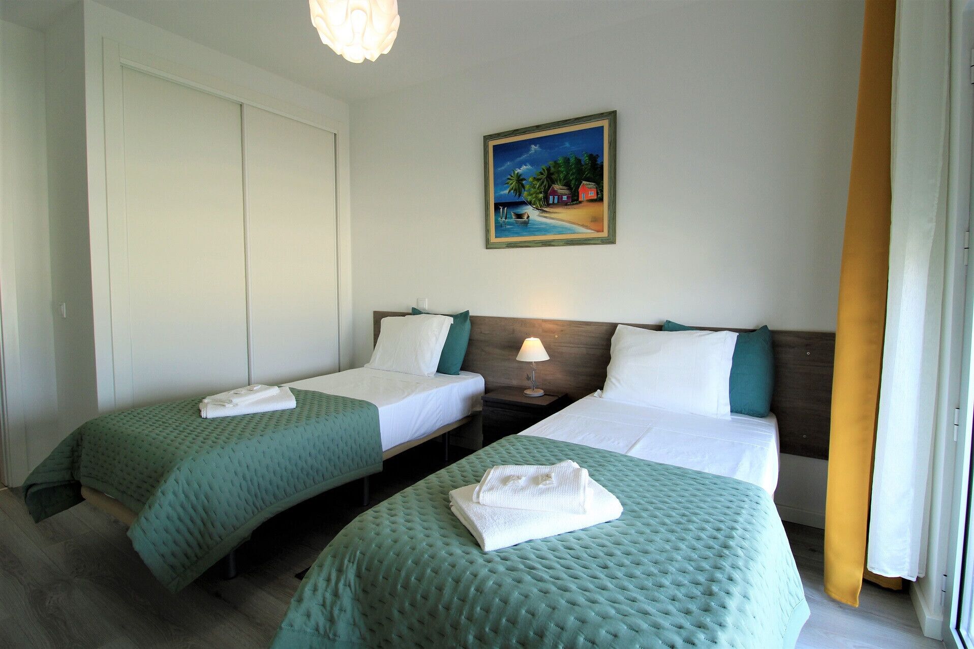 Habitación de hotel con dos camas, cada una con colcha verde y ropa de cama blanca, una mesita de noche con lámpara, un cuadro con motivo de playa en la pared y una puerta corredera.