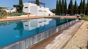 A swimming pool with a wooden deck, surrounded by cypress trees and a white building with arched windows. - Vilamoura Concept Villa With Pool (Loulé)
