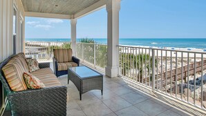 A covered outdoor area with a view of the beach, furnished with a sofa, chairs, and a table.