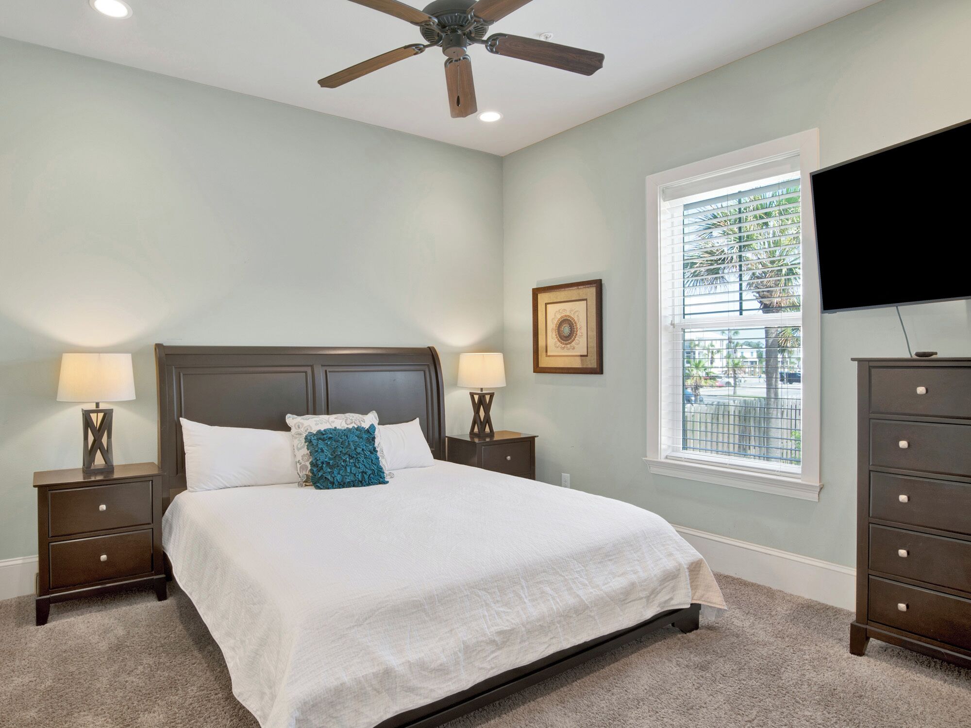 A bedroom with a bed, two bedside tables with lamps, a ceiling fan, a window with blinds, a dresser, and a TV mounted on the wall.