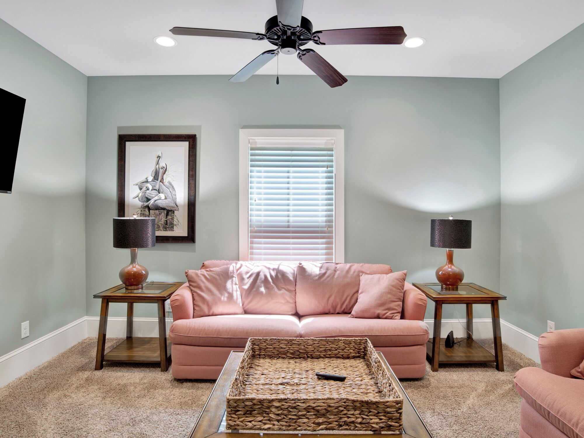 Un salon avec un canapé rose, deux tables d’appoint, un ventilateur de plafond, un tableau encadré et une fenêtre avec des stores.