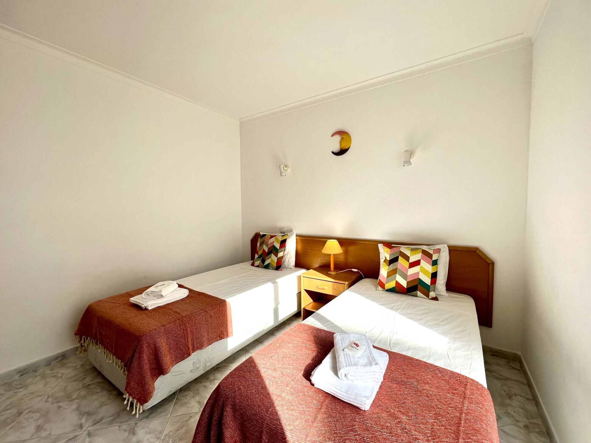 A hotel room with two single beds, a nightstand with a lamp, and a wall-mounted light fixture.