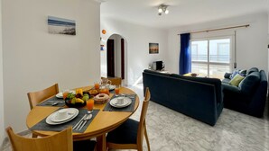 En spiseplads med et rundt bord dækket til fire personer, en blå sofa og havudsigt gennem en åben dør.