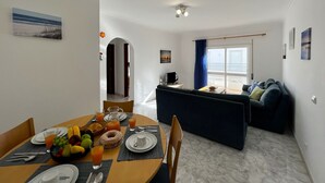 En moderne stue med en mørkeblå sofa, et spiseområde med morgenmadsbord og frugt samt juice, og en balkon med udsigt.