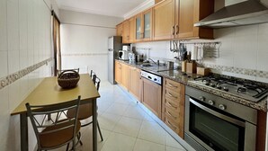 A kitchen with wooden cabinets, a stove, a dishwasher, and a dining area with chairs and a table. - Armação Pêra Typical by Homing (Silves)