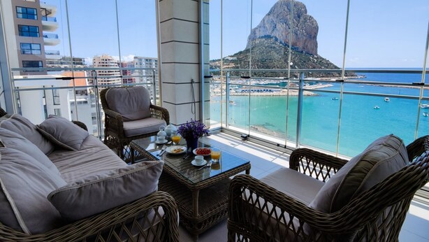 A balcony with wicker furniture, a table with breakfast items, and a view of a coastal area with buildings and a prominent rock formation. - BAUPRES luxury apartment (Calpe)