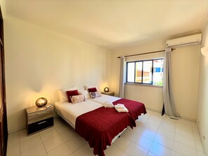 A hotel room with a bed, two bedside tables, a window with curtains, and a wall-mounted air conditioner. - Algarve Manta Rota Beach by Homing (Vila Real Santo Antonio)