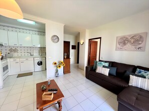 A living room with a dark brown sofa, a wooden coffee table, and a kitchen area with white cabinets and a washing machine. - Algarve Manta Rota Beach by Homing (Vila Real Santo Antonio)