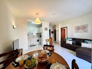 A modern living room with a dark sofa and a dining area with a round table set for breakfast. - Algarve Manta Rota Beach by Homing (Vila Real Santo Antonio)