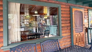 Een houten hut met een overdekte veranda, een eethoek met een tafel en stoelen, en een groot raam met gordijnen.