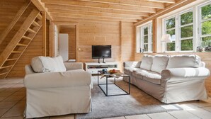Interior acolhedor de uma cabana de madeira, com uma TV, dois sofás e uma janela com vista para árvores.