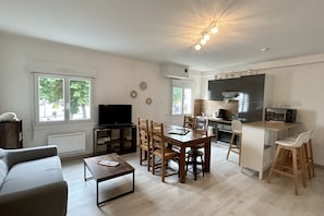 A modern living room with a sofa, coffee table, TV, dining table with chairs, kitchen area with appliances, and a large window with a view of trees.