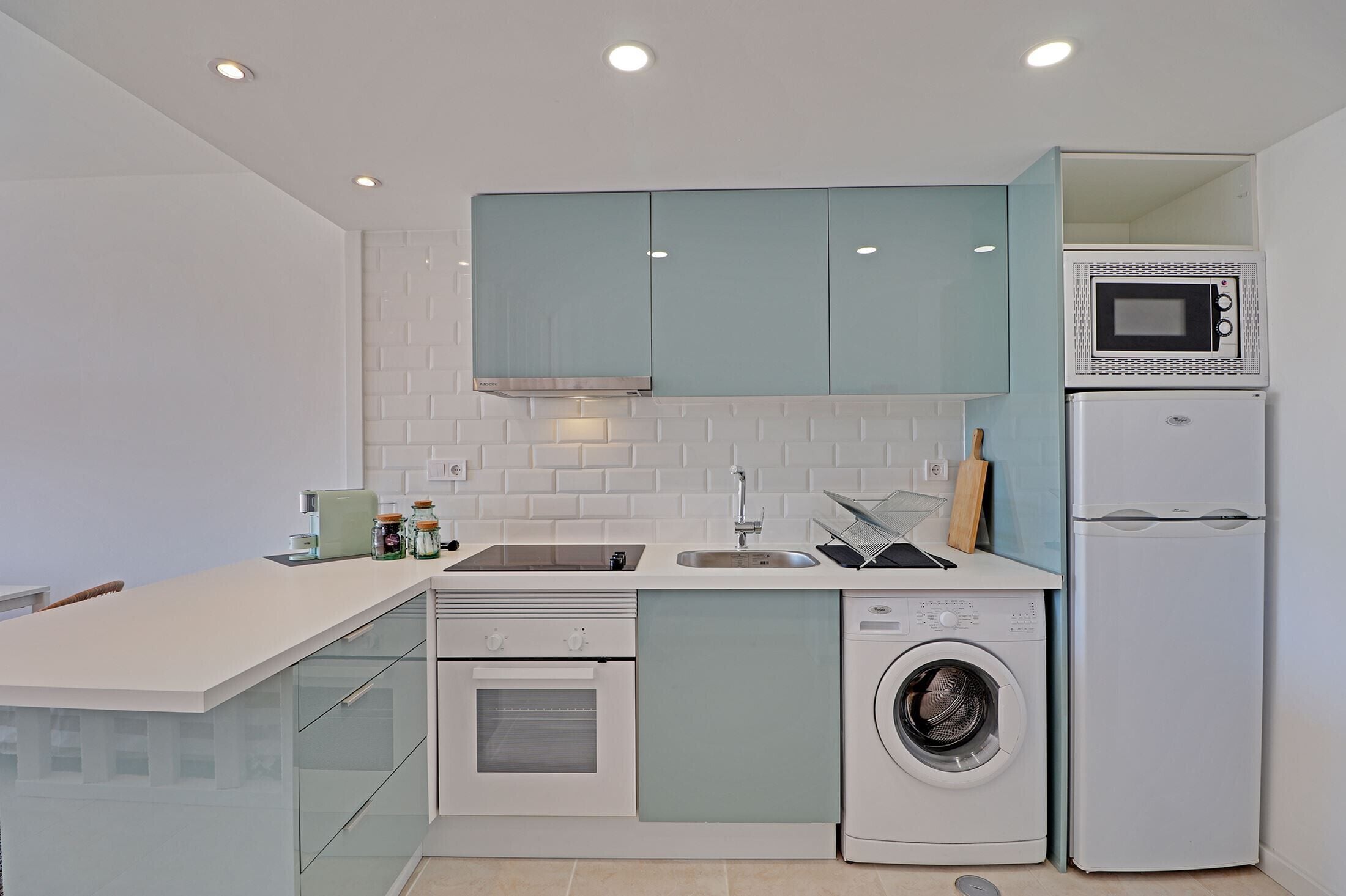 A compact kitchen with a built-in oven, microwave, and washing machine, featuring a modern design with light blue cabinets and white countertops.