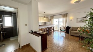 Een ruime woonkamer met houten vloer, een eettafel met stoelen, een bank en een klein keukenhoekje met raam.
