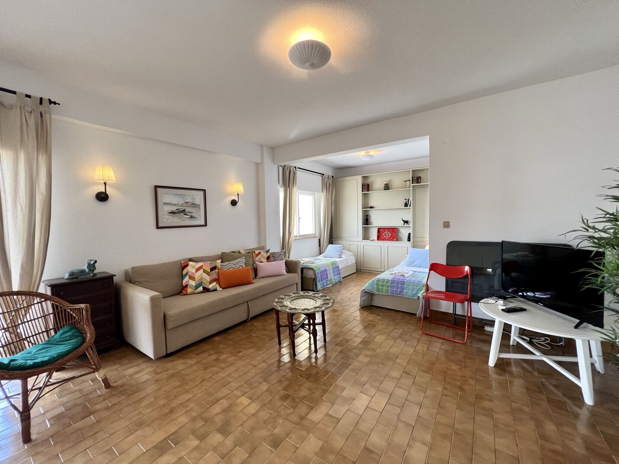 A well-lit living room with a sofa, two beds, a TV, and a dining area.