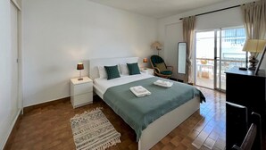 Een hotelkamer met een bed, nachtkastje, stoel, spiegel en een balkon met uitzicht.