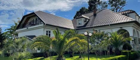 A two-story house with a balcony, surrounded by well-maintained gardens and a clear sky.