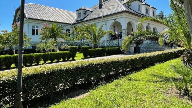 A large, two-story building with a white facade and a grey roof, surrounded by well-maintained hedges and palm trees. - Hotel Laguna (Ureki)
