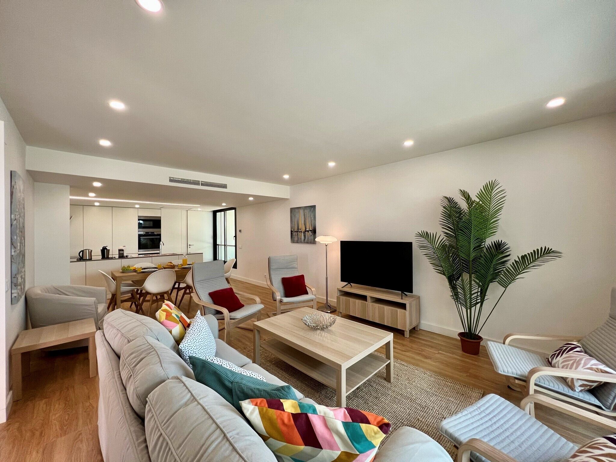 A modern living room with a sofa, chairs, a coffee table, a TV, and a dining area with a kitchen in the background.