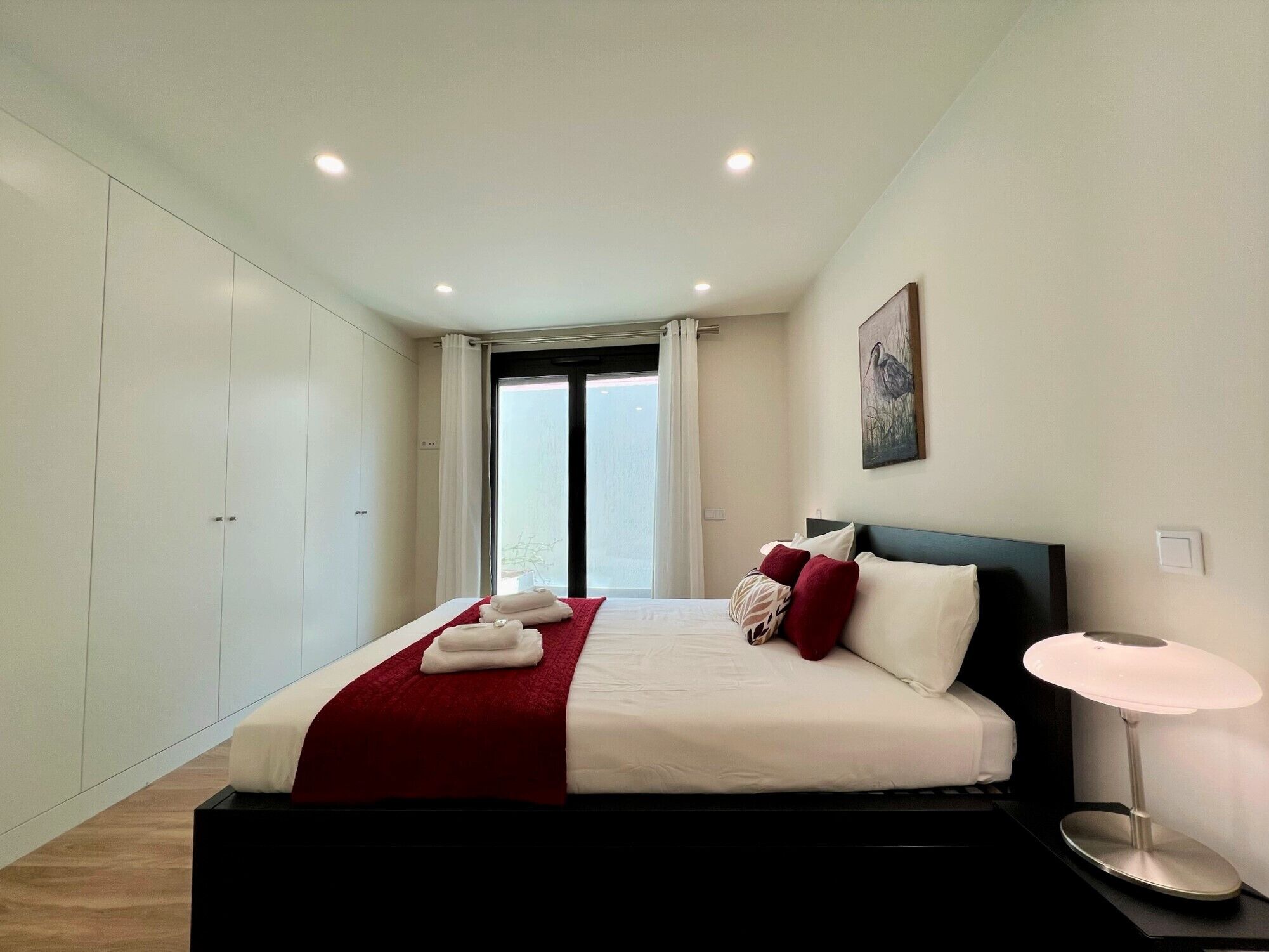 A modern bedroom with a large bed, a bedside table with a lamp, a window with curtains, and a wall-mounted artwork.