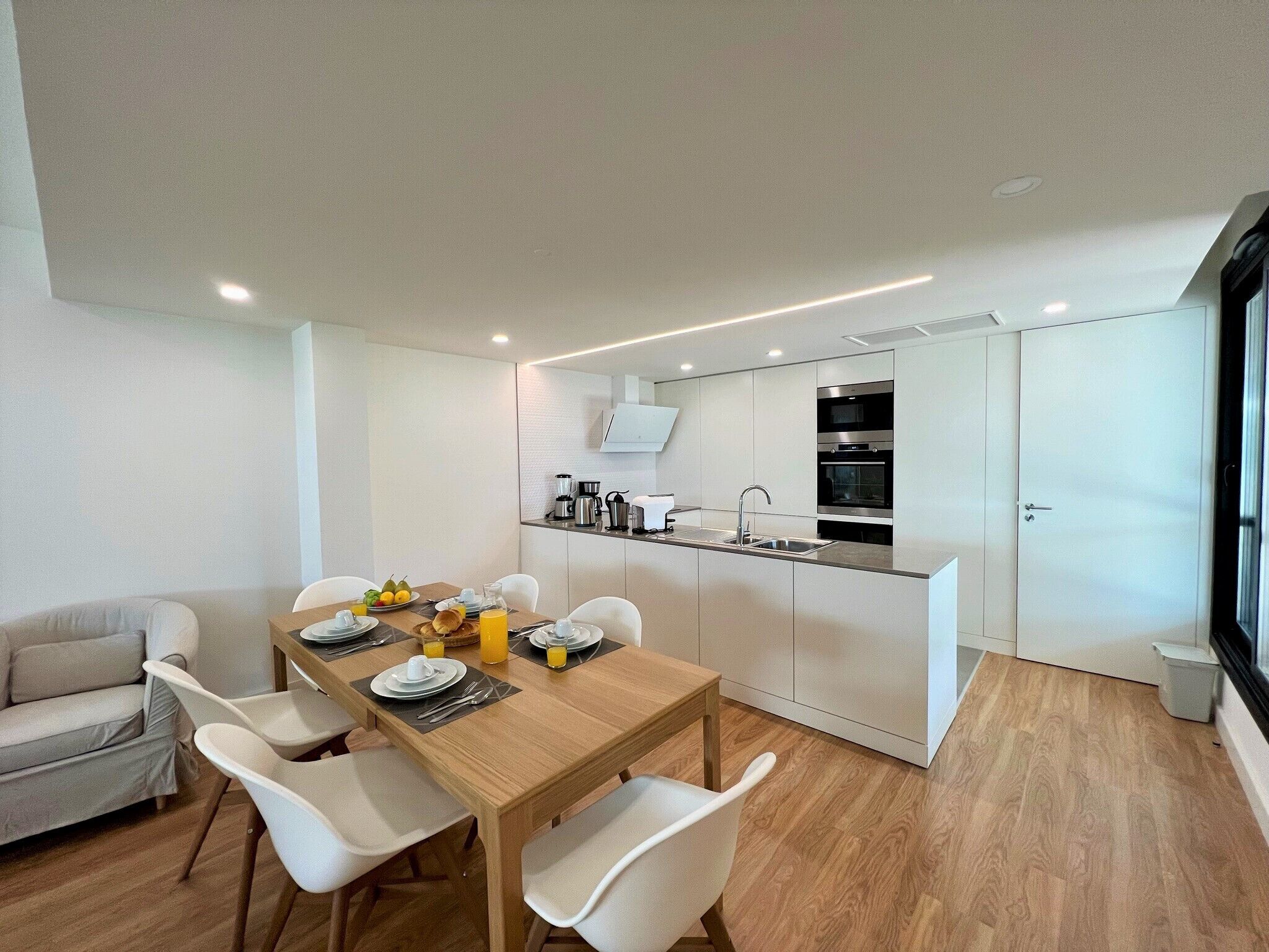 A modern kitchen with built-in appliances, a dining area with a wooden table and white chairs, and a large window providing natural light.