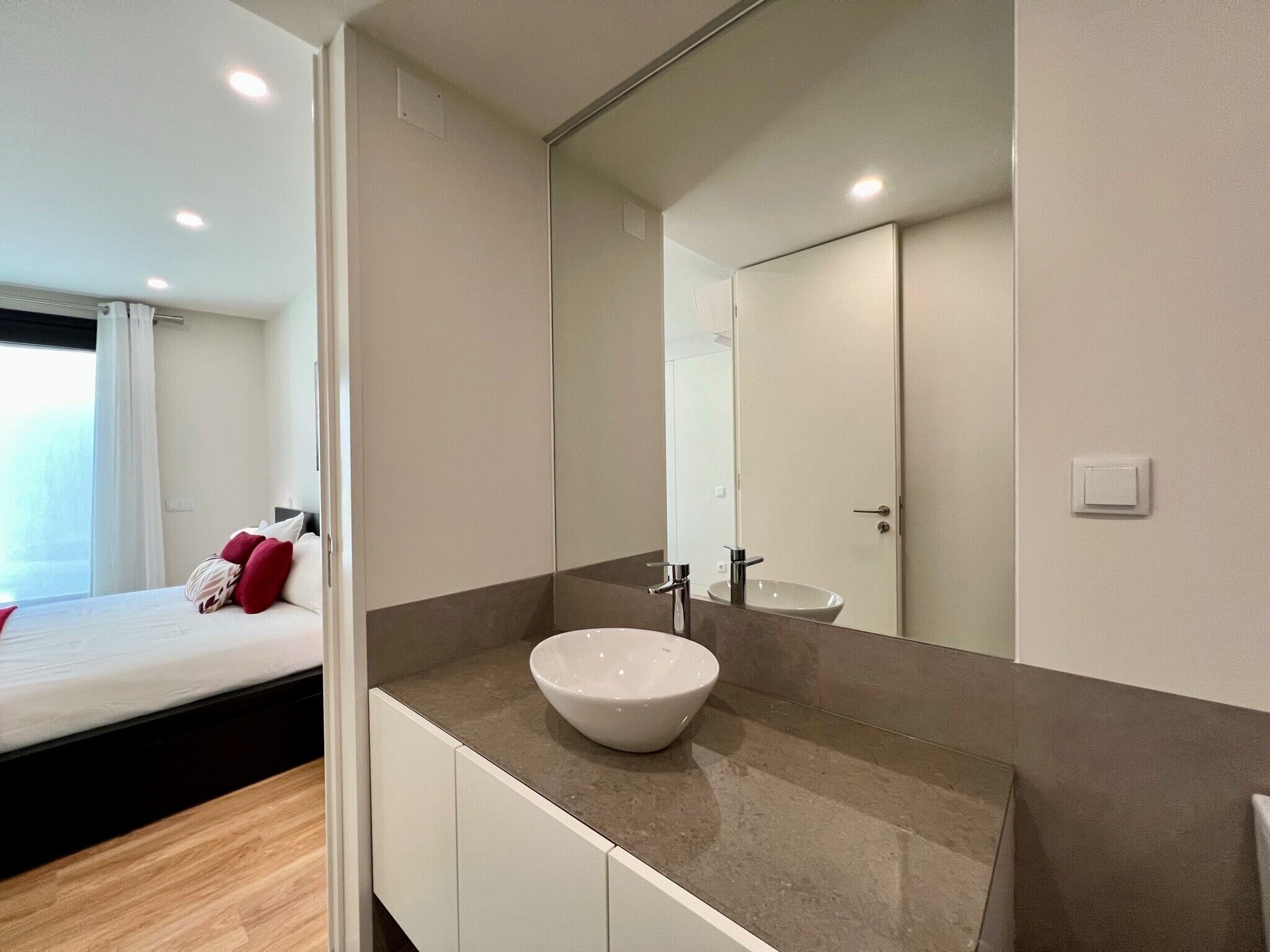 A modern bathroom with a large mirror, a white sink, and a wooden floor.