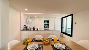 A modern dining area with a wooden table set for two, a kitchen in the background, and a large window providing natural light. - Faro Design 6 by Homing (Faro)