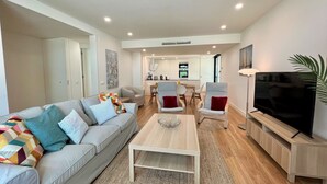 A modern living room with a sofa, coffee table, and TV stand. There is a dining area with chairs and a kitchen in the background. - Faro Design 6 by Homing (Faro)