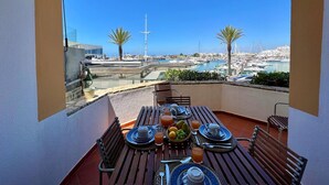 Mesa en balcón dispuesta con alimentos para el desayuno, con vista a un puerto deportivo con barcos y palmeras.