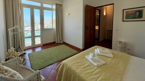 Un cuarto de hotel con una cama, una alfombra verde, una silla de mimbre, vista al mar a través de ventanas amplias y un cuadro en la pared.