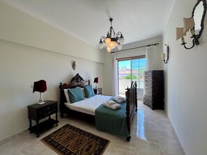 Ein Schlafzimmer mit Bett, Nachttischen, einer Chandelüster, einem Schminktisch und Blick ins Freie.