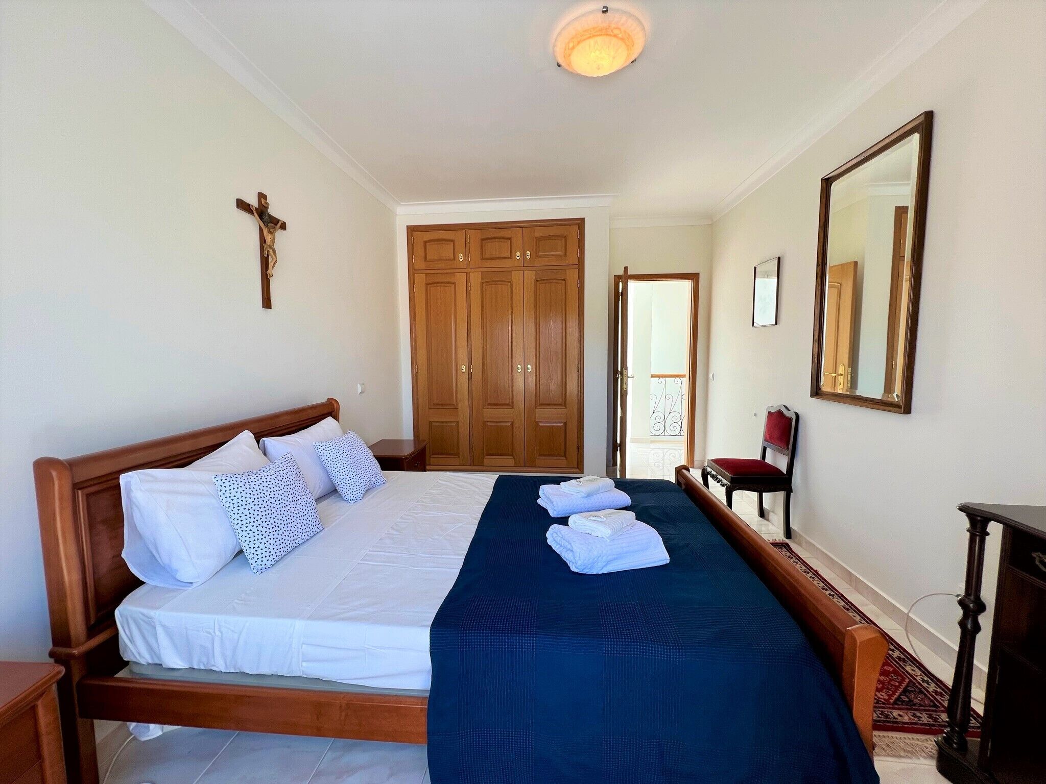 A bedroom with a wooden bed, a cross on the wall, a wardrobe, and a mirror.