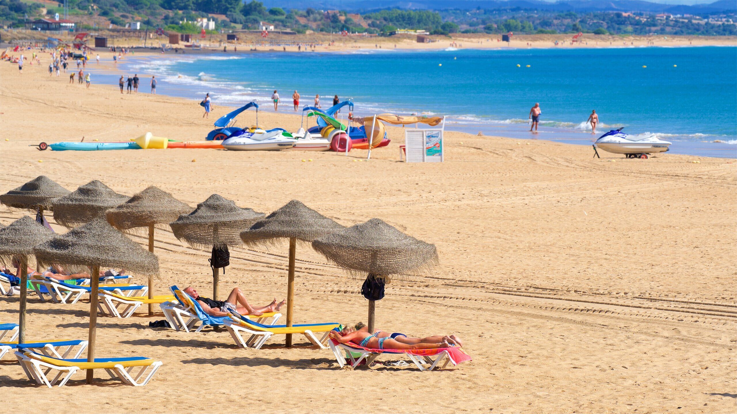 A beach with sun loungers and umbrellas, a lifeguard tower, and boats in the background.