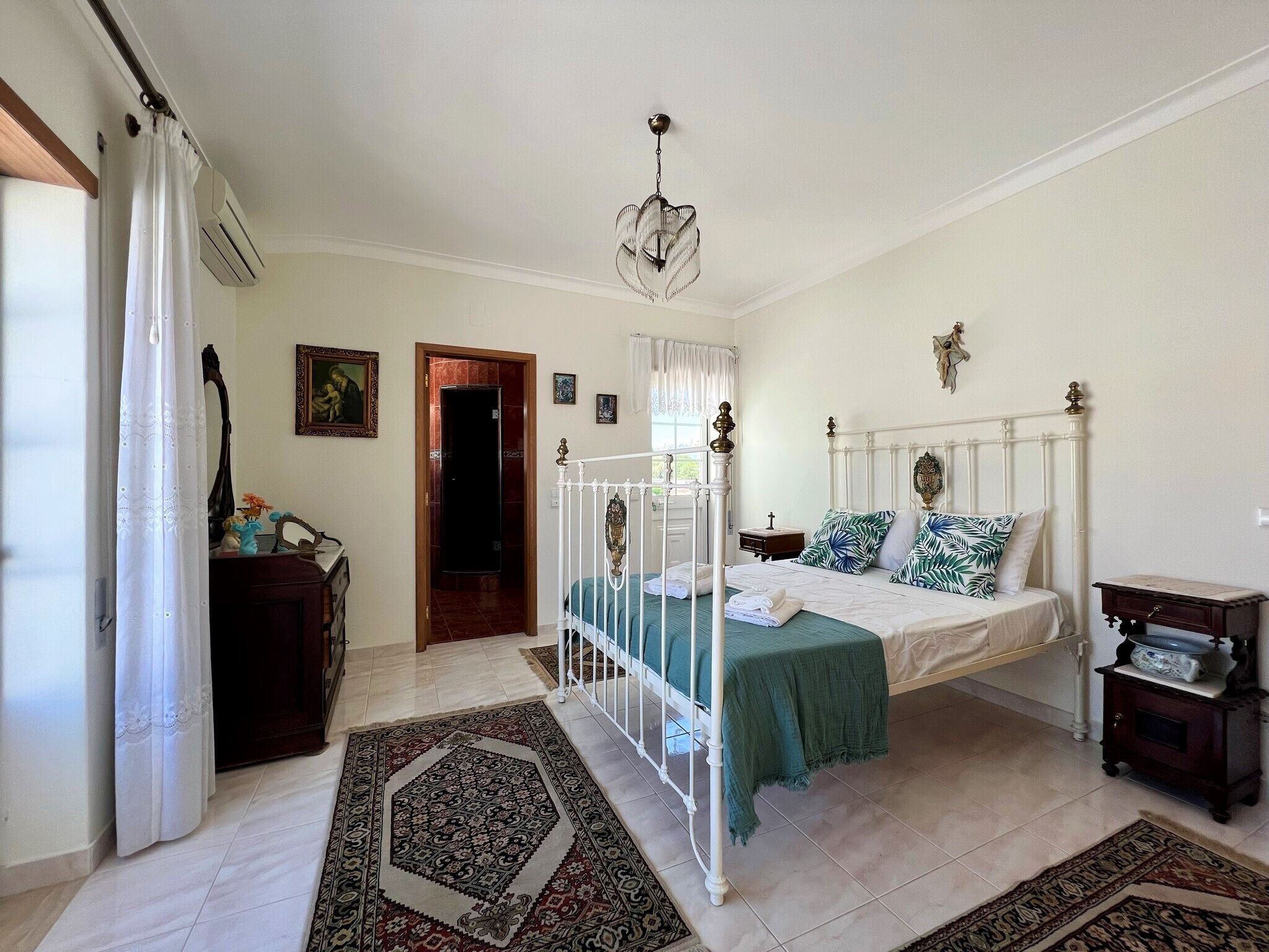 Ein Schlafzimmer mit Bett, Nachttisch, Kommode, einer Pendelleuchte und einer Tür zu einem anderen Raum.
