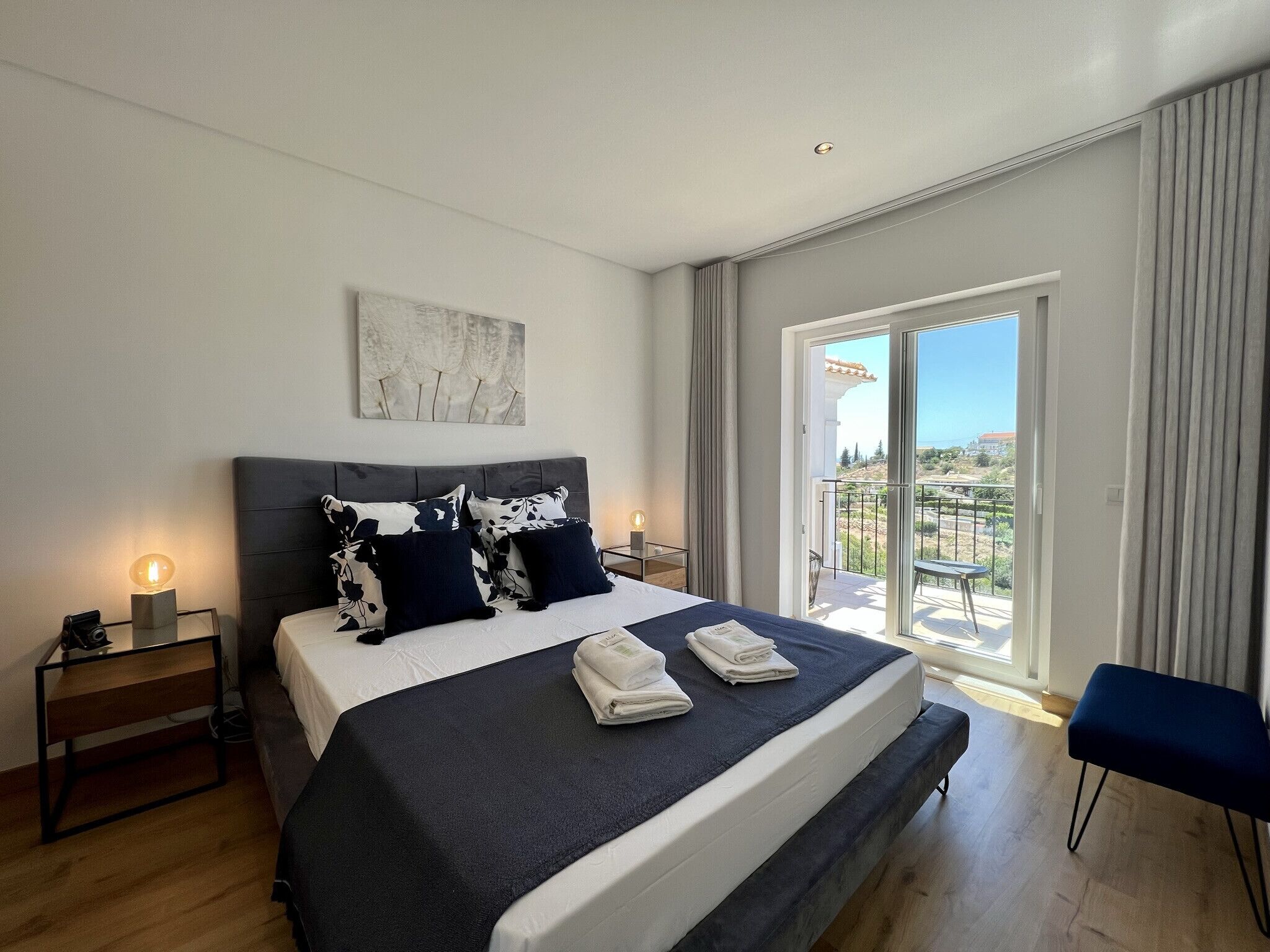A modern bedroom with a large bed, two bedside tables, a nightstand, a chair, and a balcony with a view.
