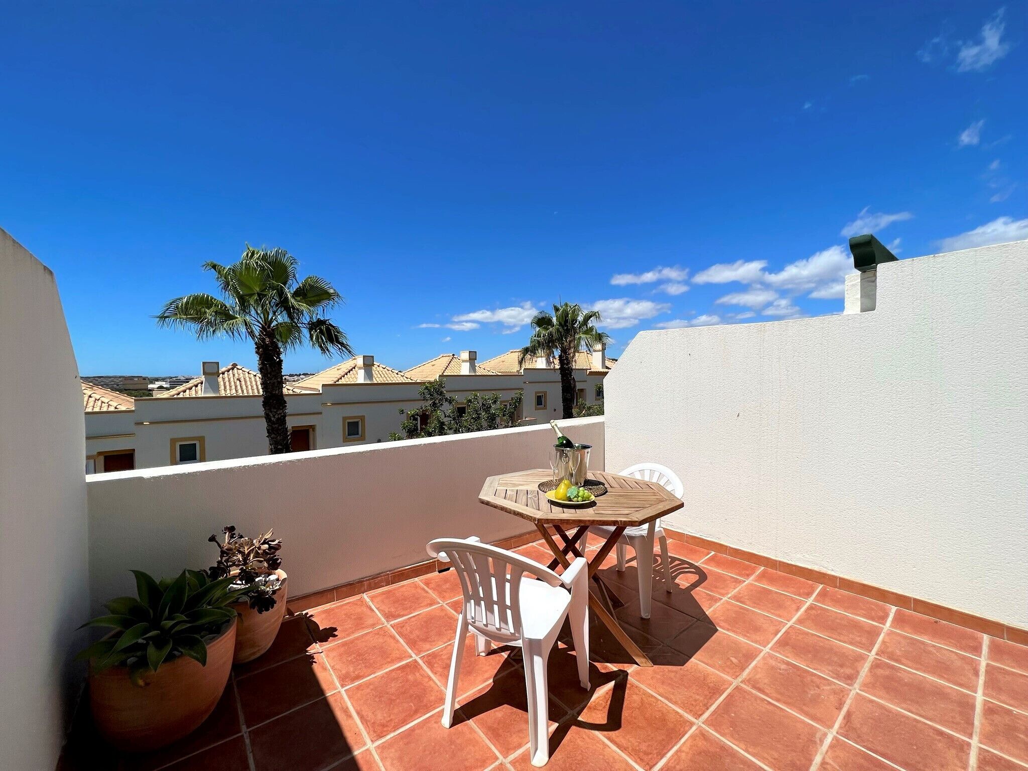 A terrace with a table and chairs, potted plants, and a view of buildings and palm trees.