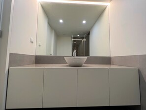 A modern bathroom with a large mirror, a white sink, and a shower visible in the background. - Faro Design 3 by Homing (Faro)