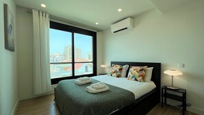 Een moderne hotelkamer met een groot bed, twee bedlampjes, een raam met uitzicht op gebouwen en een airconditioner.