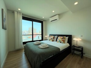 Ein modernes Hotelzimmer mit einem großen Bett, zwei Nachttischlampen, einem Fenster mit Blick auf Gebäude und einer Klimaanlage.