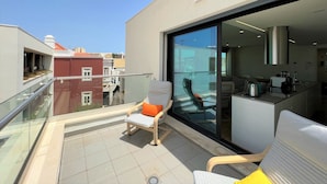 Een balkon met een stoel en tafel, een glazen deur die leidt naar een keuken, en uitzicht op omliggende gebouwen.