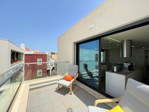Ein Balkon mit Stuhl und Tisch, eine Glastür zu einer Küche und Blick auf benachbarte Gebäude.