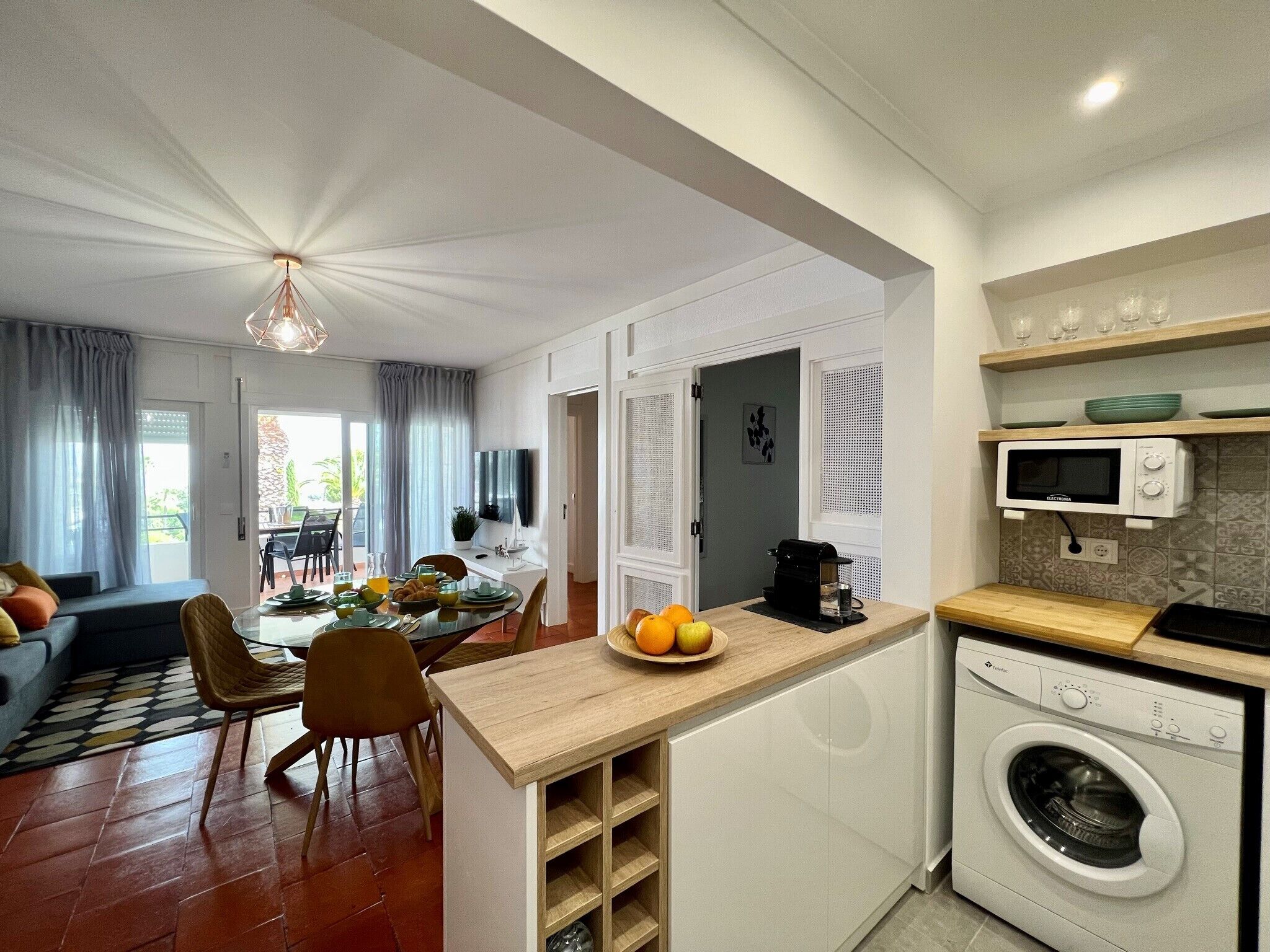 Cozinha moderna com máquina de lavar embutida, área de refeições com mesa e cadeiras, e sala de estar com sofá e televisão.