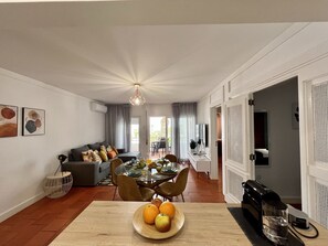 A modern living room with a dining area, featuring a wooden table with fruit, a sofa with cushions, and a coffee machine. - Armação DE Pêra Brightness 2 With Pool (Lagoa)