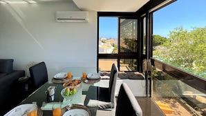 A modern dining area with a glass table, chairs, and a view of the cityscape through large windows. - Albufeira Central 2 by Homing (Albufeira)