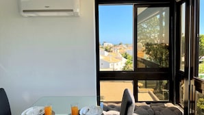 A dining area with a table set for breakfast, a large window with a view of buildings and trees, and an air conditioning unit on the wall. - Albufeira Central 2 by Homing (Albufeira)
