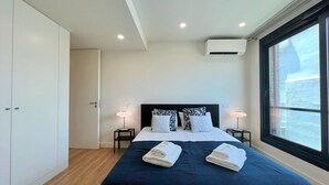 A modern hotel room with a large bed, two bedside tables with lamps, a wardrobe, an air conditioning unit, and a window with a view. - Faro Design 1 by Homing (Faro)
