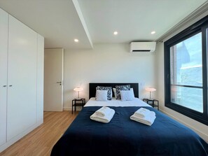 Ein modernes Hotelzimmer mit einem großen Bett, zwei Nachttischen mit Lampen, einem Kleiderschrank, einer Klimaanlage und einem Fenster mit Blick.