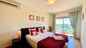 A hotel room with a large bed, a nightstand, a lamp, a window with curtains, and a view of the city. - Albufeira Modern 2 by Homing (Albufeira)